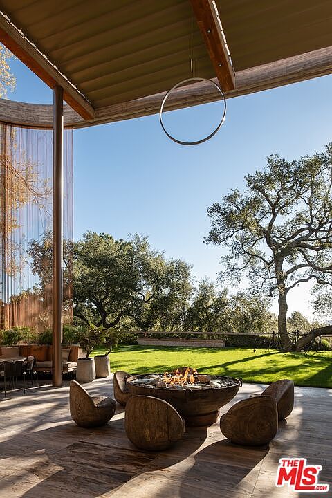 This image showcases a luxurious outdoor patio area, featuring a modern fire pit surrounded by unique wooden chairs. The patio is constructed with large, light-colored stone tiles, and the space is partially covered by a contemporary roof structure with a circular light fixture. The backdrop includes a well-manicured lawn and mature trees, creating a serene and inviting atmosphere.