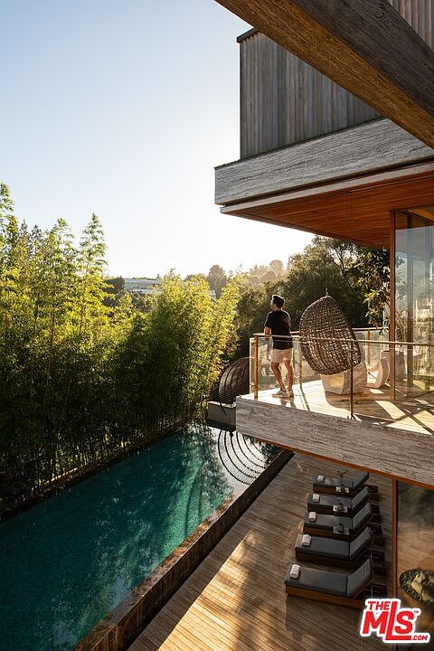 This image showcases a modern home's exterior, highlighting a sleek, narrow pool adjacent to a wooden deck. The deck features stylish lounge chairs, and a balcony above offers a view of the lush greenery surrounding the property. The architectural design emphasizes clean lines and natural materials, creating a luxurious and serene atmosphere.