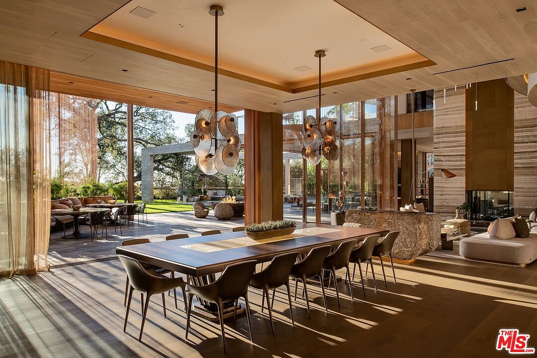 This is a spacious dining room with floor-to-ceiling windows offering views of the outdoor area. A long, dark wood dining table with modern chairs sits beneath two unique, multi-tiered chandeliers. The room features hardwood floors and a neutral color palette, creating an elegant and inviting atmosphere.