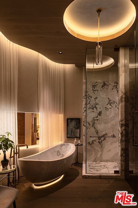 This is a luxurious primary bathroom featuring a freestanding marble bathtub with underlighting, a glass-enclosed shower with marble walls, and elegant sheer curtains. The room is illuminated by a unique circular ceiling light fixture, creating a spa-like ambiance. The overall design is modern and sophisticated, emphasizing high-end materials and a serene atmosphere.