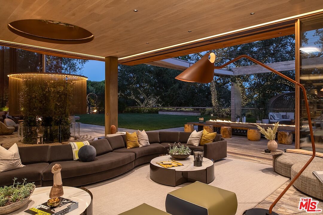 This is a luxurious living room featuring a curved brown sofa, a round marble-topped coffee table, and a unique overhead lighting fixture. The room opens to an outdoor space with a fire pit and lush greenery, creating a seamless indoor-outdoor living experience. A large, modern floor lamp adds to the room's sophisticated ambiance.