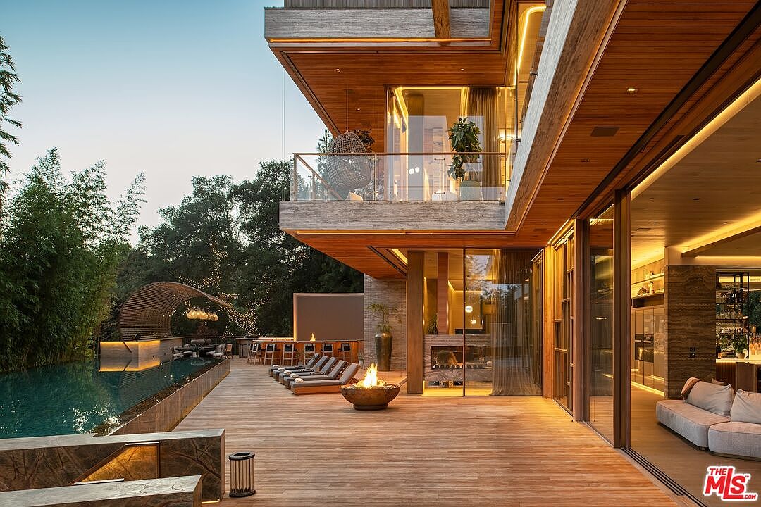 This image showcases a luxurious outdoor living space with a modern architectural design. The expansive wooden deck features a fire pit, lounge chairs, and a bar area, leading to a stunning pool and a unique bamboo-covered seating area. The multi-level structure includes a balcony with glass railings and a hanging chair, seamlessly blending indoor and outdoor living.