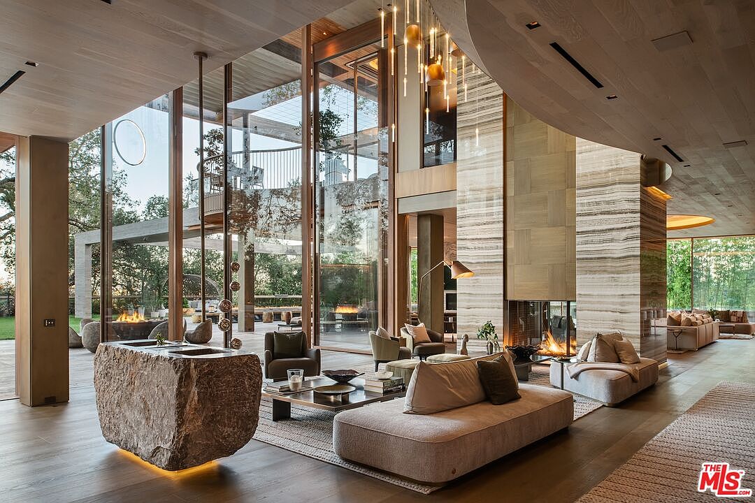 This is a luxurious living room featuring floor-to-ceiling windows that offer views of the surrounding landscape. The room is furnished with modern sofas, a stone coffee table, and a unique stone block feature. The high ceilings and natural light create an open and airy atmosphere, enhancing the sense of spaciousness and elegance.