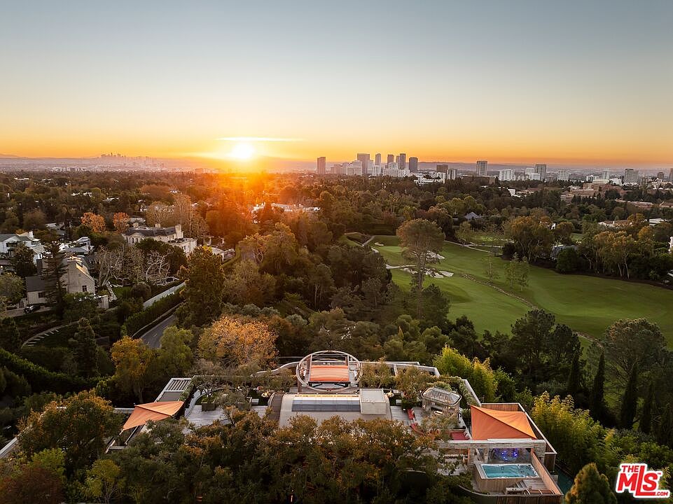 This aerial view showcases a luxurious estate at sunset, featuring a modern rooftop deck with unique architectural elements, a swimming pool, and meticulously landscaped grounds. The property overlooks a lush green golf course and offers panoramic city views, creating an impression of exclusivity and high-end living.