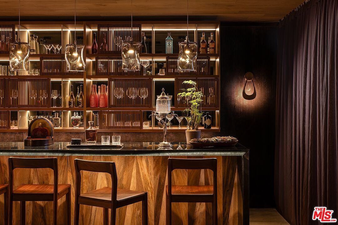 This image showcases a sophisticated wine cellar featuring a custom-built wooden shelving system with integrated lighting, displaying an array of bottles and glassware. The bar area includes a dark countertop and wooden bar stools, complemented by unique pendant lighting and a decorative wall sconce. The overall ambiance is luxurious and inviting, perfect for entertaining.