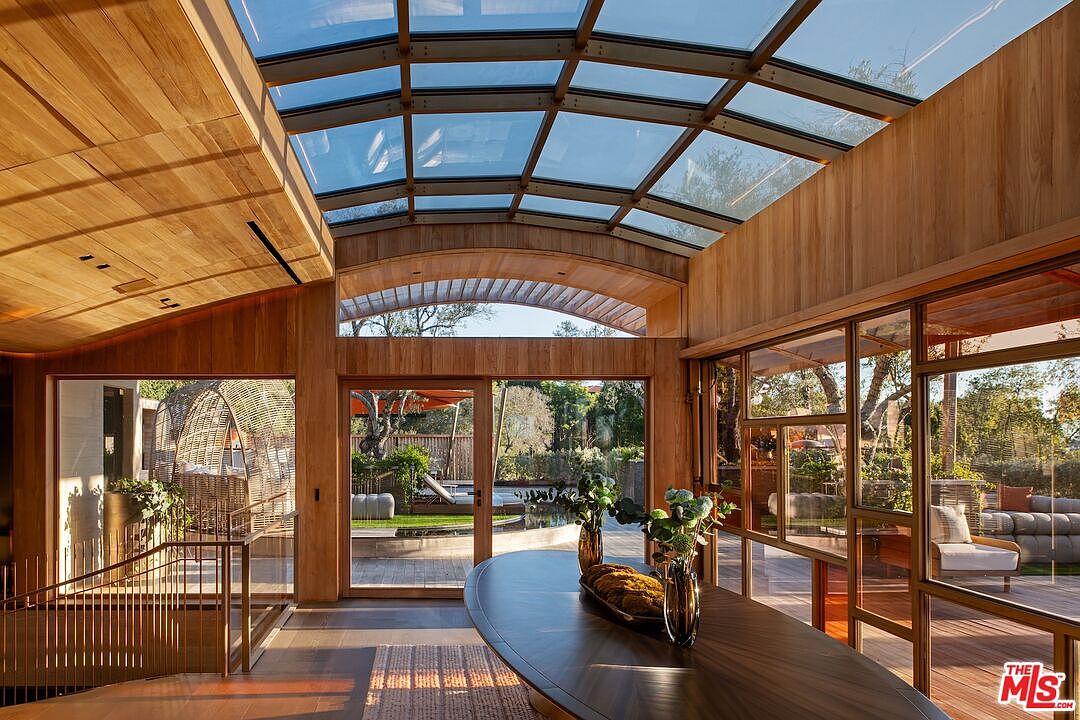 This interior shot showcases a dining room with a modern, nature-inspired design. The room features wood paneling, a glass ceiling, and large windows that offer views of the outdoor landscape. A dark wood dining table is the focal point, complemented by natural light and a seamless transition to the exterior spaces.