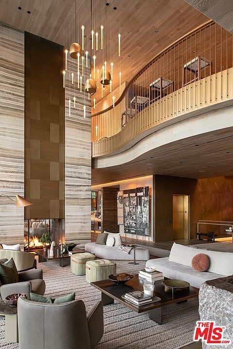 This is a luxurious living room featuring a soaring two-story space with a striking fireplace wall made of stone and wood. The room is furnished with modern sofas, armchairs, and a coffee table, all arranged on a large area rug. A unique chandelier hangs from the ceiling, adding to the room's grandeur, and a balcony overlooks the living area.