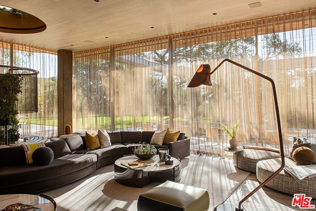 This is a luxurious living room featuring a curved sofa, a round marble-topped coffee table, and floor-to-ceiling windows with sheer curtains that offer a view of the lush greenery outside. A modern arc floor lamp adds a stylish touch, and the overall design exudes sophistication and comfort. The room is bathed in natural light, creating a warm and inviting atmosphere.