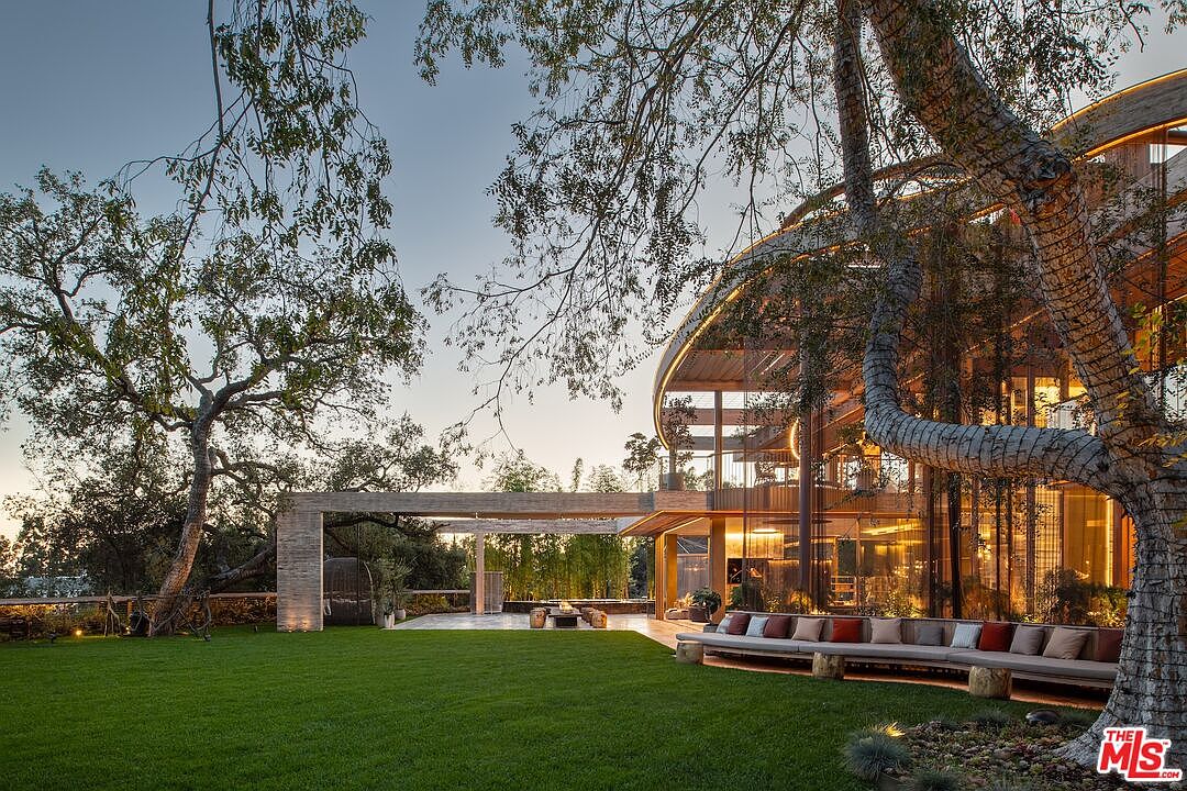 This image showcases the expansive yard and garden of a luxury property, featuring a meticulously maintained lawn, mature trees, and a modern architectural structure. A covered outdoor seating area with a fire pit and a long bench with cushions provide ample space for relaxation and entertainment, highlighting the property's focus on outdoor living and sophisticated design.