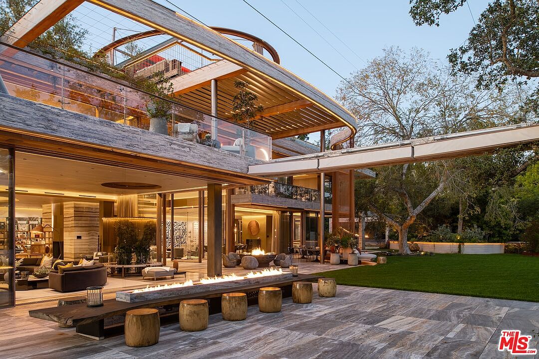 This exterior shot showcases a luxurious patio area with a modern fire feature, wooden stools, and sleek stone flooring. The patio seamlessly connects to the interior living space through large glass doors, offering an open and airy feel. The architectural design is contemporary, with clean lines and natural materials, creating an inviting outdoor living space.