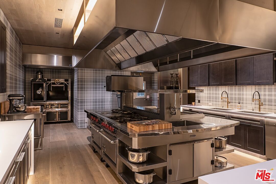 This is a professional-grade kitchen featuring stainless steel appliances, a large central island with a cooktop, and a commercial-style range hood. The walls are covered in a plaid pattern, and the cabinets are a dark wood tone. The overall impression is one of high-end functionality and sophisticated design.