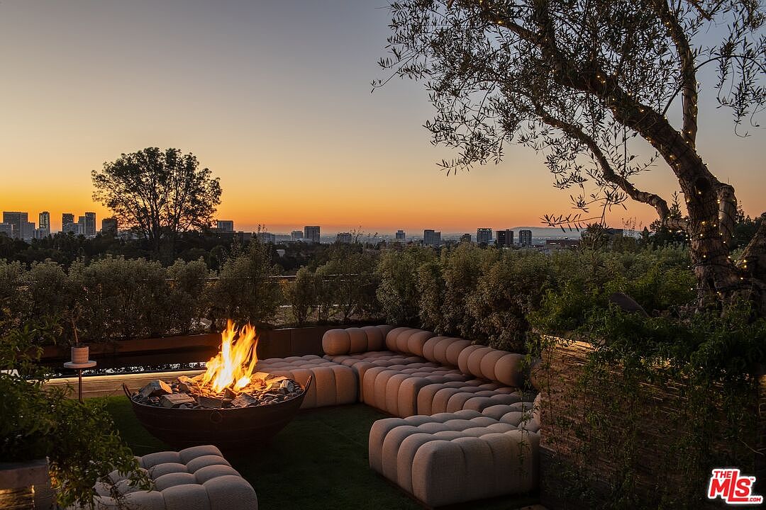This image showcases a luxurious outdoor patio area at sunset, featuring a modern, rounded sectional sofa arranged around a fire pit. Lush greenery surrounds the seating area, providing privacy and a sense of tranquility, while the city skyline is visible in the distance, adding a touch of urban sophistication. The overall impression is one of upscale relaxation and entertainment.
