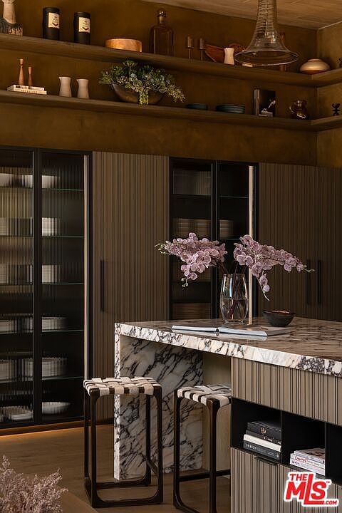 This interior shot showcases a modern kitchen with sleek, vertically paneled cabinetry and a striking marble countertop island. The kitchen features glass-front cabinets displaying dishware, complemented by stylish bar stools and decorative floral arrangements. The warm lighting and sophisticated design create an inviting and luxurious atmosphere.