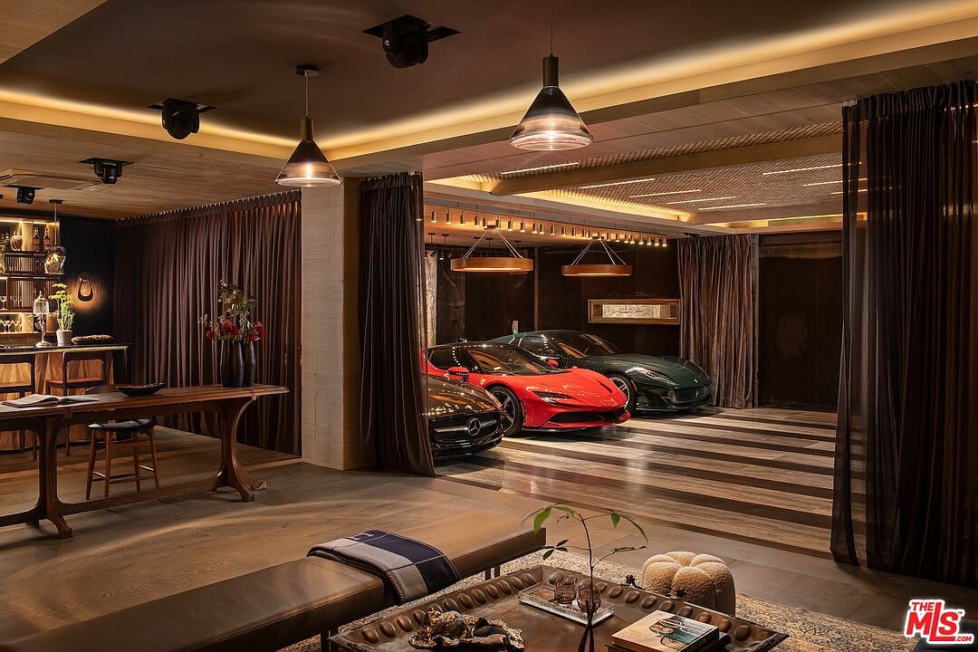This interior shot showcases a luxurious living space that doubles as a garage, featuring a curated collection of cars as a focal point. The room is elegantly designed with warm lighting, rich wood tones, and sophisticated furnishings, creating a unique blend of automotive display and comfortable living. Dark curtains add a dramatic flair to the space.