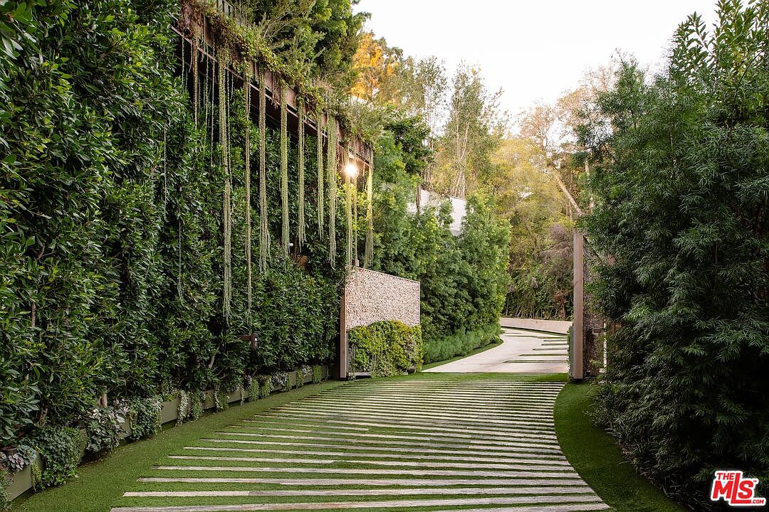 This image showcases a grand entryway featuring a unique driveway design with alternating concrete slabs and grass, leading to a gated entrance. Lush greenery and vertical gardens adorn the walls, creating a private and luxurious feel. A stone accent wall adds texture and visual interest, enhancing the overall curb appeal.