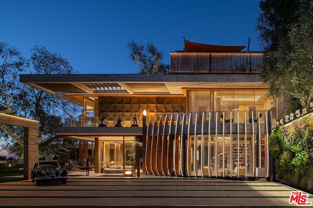 This is a stunning front exterior view of a modern luxury home at dusk. The architectural design features clean lines, a multi-level structure with a rooftop deck, and extensive use of glass and wood. The property includes a meticulously landscaped yard and a classic car parked in the driveway, adding to the overall impression of sophistication and high-end living.