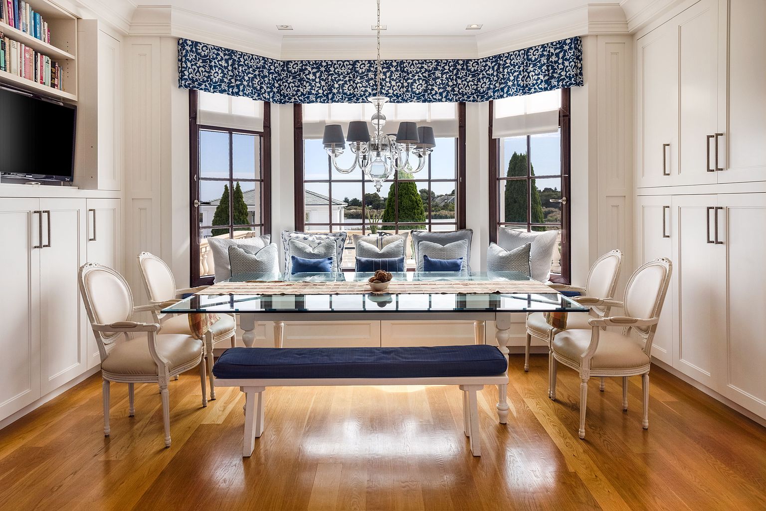 This is an interior shot of a dining room featuring a glass-top table with white legs, surrounded by elegant white chairs and a bench with a blue cushion. The room is well-lit by a chandelier and natural light streaming through a bay window with a blue patterned valance, offering a view of the outdoors. Built-in white cabinets flank the dining area, adding to the room's sophisticated and organized feel.