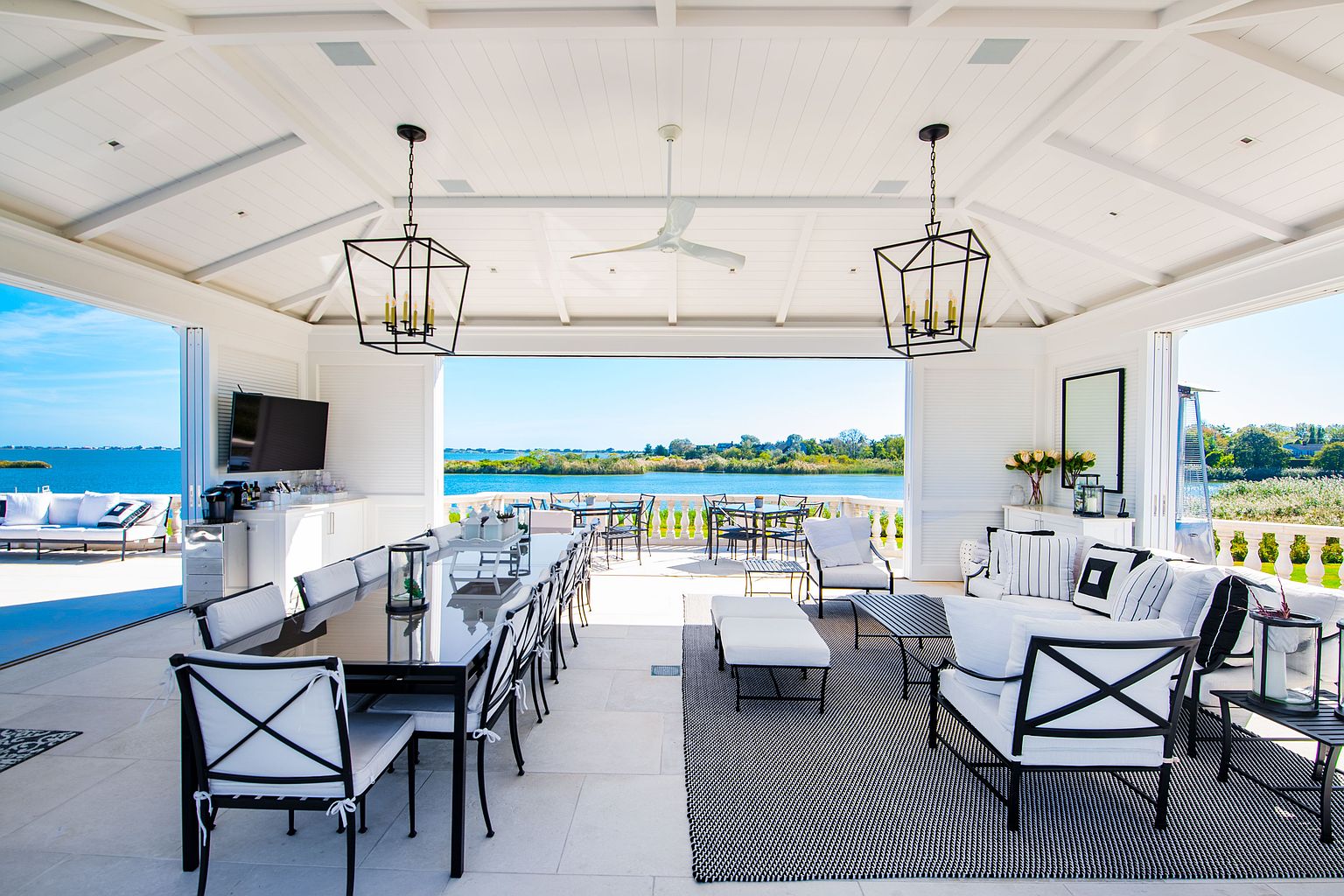 This image showcases a luxurious outdoor living space, featuring a covered patio or balcony with elegant black and white furniture, including a long dining table and comfortable seating areas. The space is open to a stunning waterfront view, creating a seamless indoor-outdoor transition. The architectural details, such as the white vaulted ceiling and stylish light fixtures, add to the overall upscale ambiance.