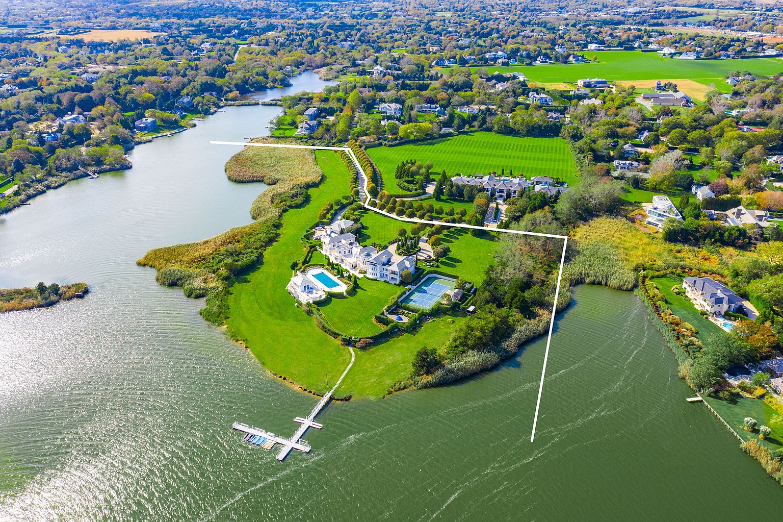 This aerial shot showcases a luxurious waterfront estate with sprawling green lawns, a large main house, a pool, and a tennis court. The property is bordered by a serene lake and lush landscaping, offering privacy and stunning views. A long dock extends into the water, providing access for boating and other water activities.