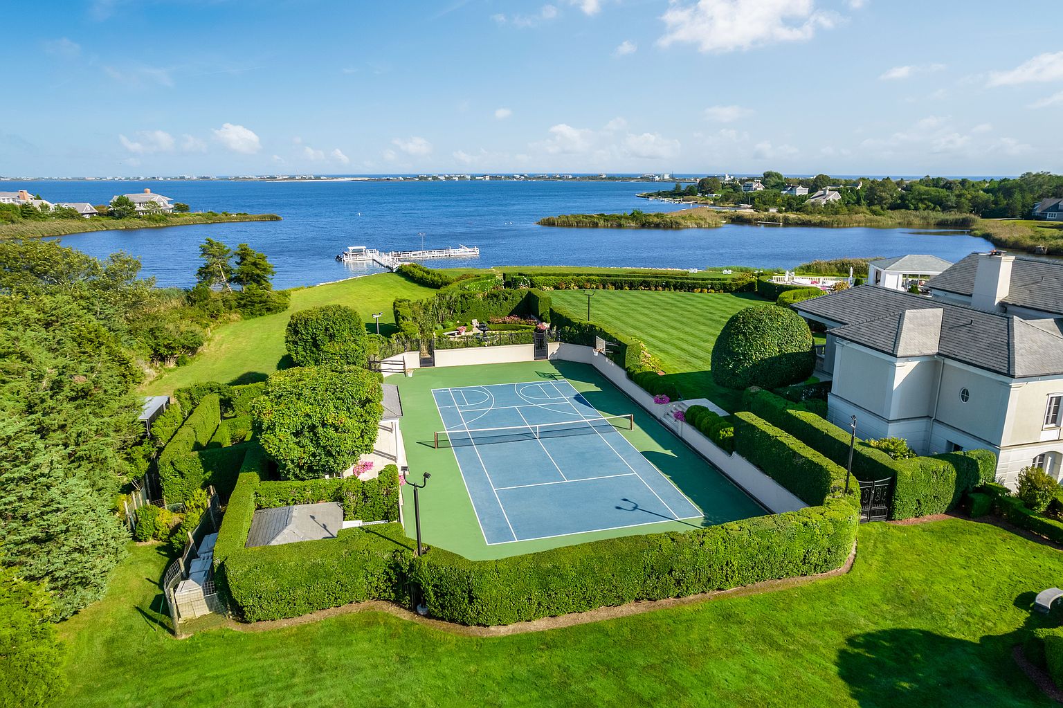 This aerial view showcases a luxurious estate with meticulously manicured grounds, including a tennis court enclosed by hedges. The property features a large house with a gray roof, expansive lawns, and waterfront access with a dock. The overall impression is one of opulence and privacy.