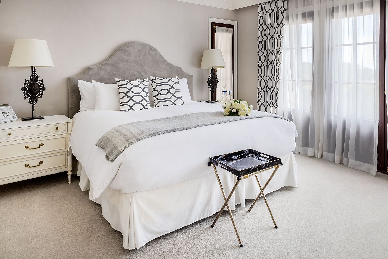 This is a well-appointed primary bedroom featuring a plush bed with a grey headboard, white linens, and decorative pillows. A bedside table with a lamp and a small tray table add to the room's functionality and style. The room is bright and inviting, with natural light streaming through the windows, creating a serene and comfortable atmosphere.