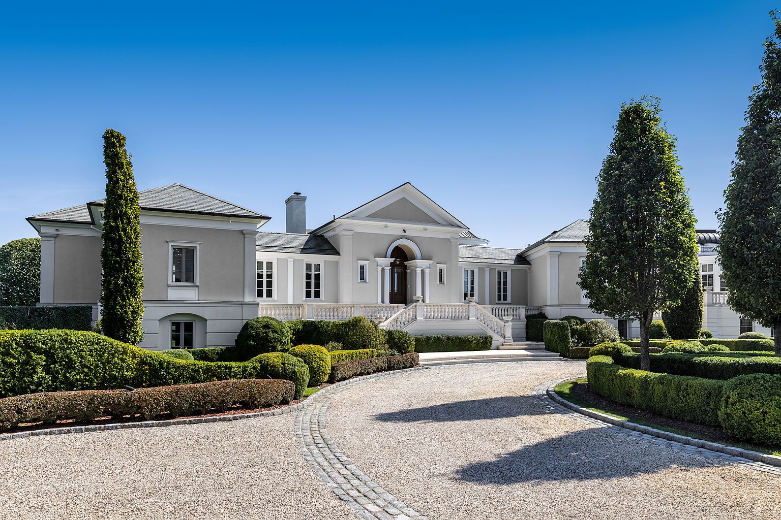 This is a front view of a grand, classically styled home with a symmetrical facade. The house features a light gray exterior, white trim, and a prominent entrance with columns and a balustraded staircase. Manicured landscaping, including shaped hedges and gravel driveway, enhances the property's curb appeal, creating an impression of luxury and meticulous care.