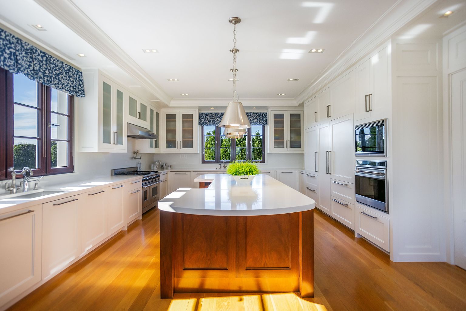 This is a bright and spacious kitchen featuring white cabinetry, stainless steel appliances, and a large wooden island with a white countertop. Natural light floods the room through multiple windows, complemented by recessed lighting and a pendant light above the island. The hardwood floors add warmth to the space, creating an inviting atmosphere.