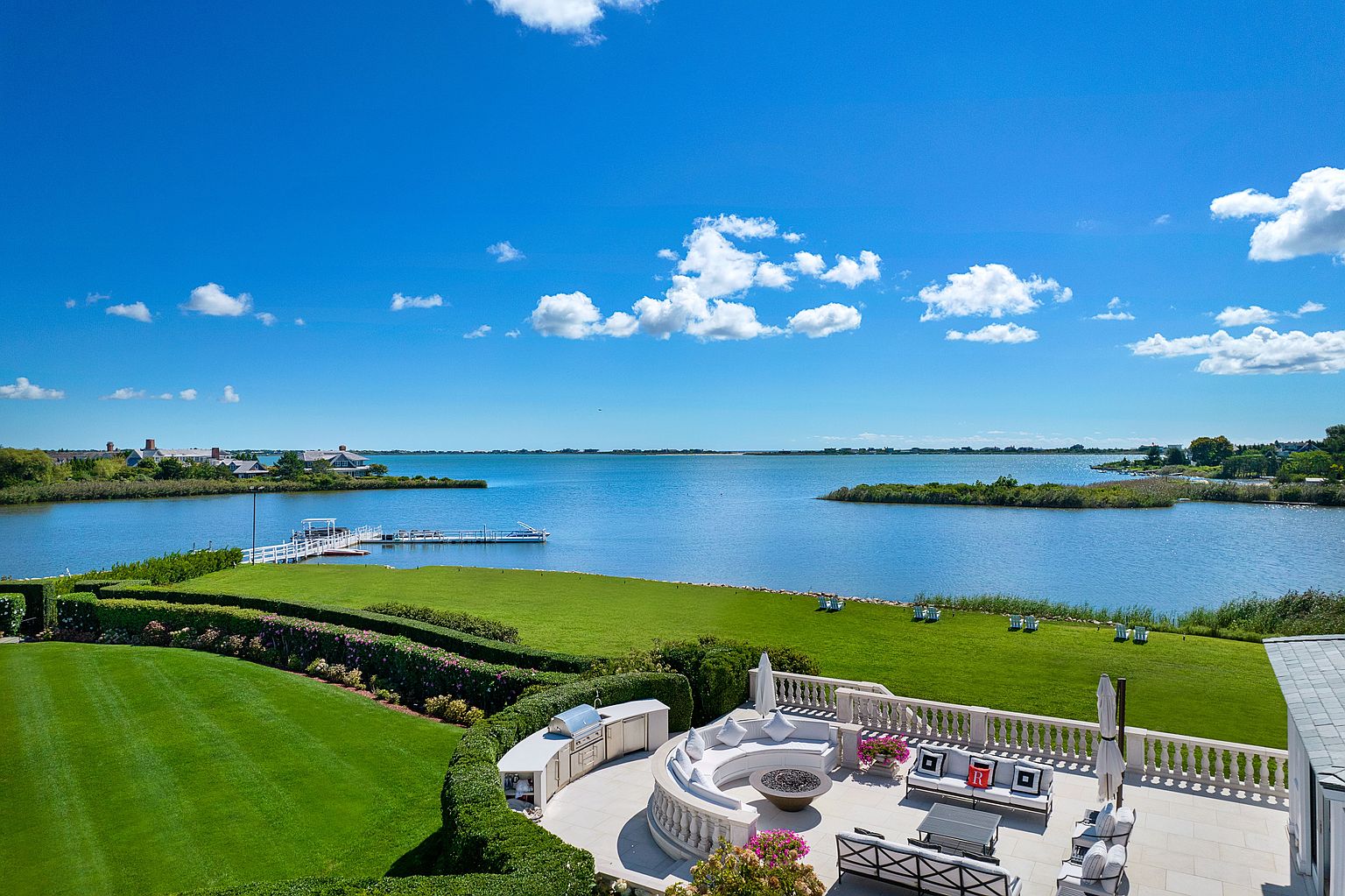 This image showcases a luxurious outdoor patio area with a stunning waterfront view. The patio features a circular seating area with a fire pit, elegant outdoor furniture, and a built-in grilling station. Manicured lawns and hedges surround the patio, leading to the water's edge, creating a serene and upscale ambiance.