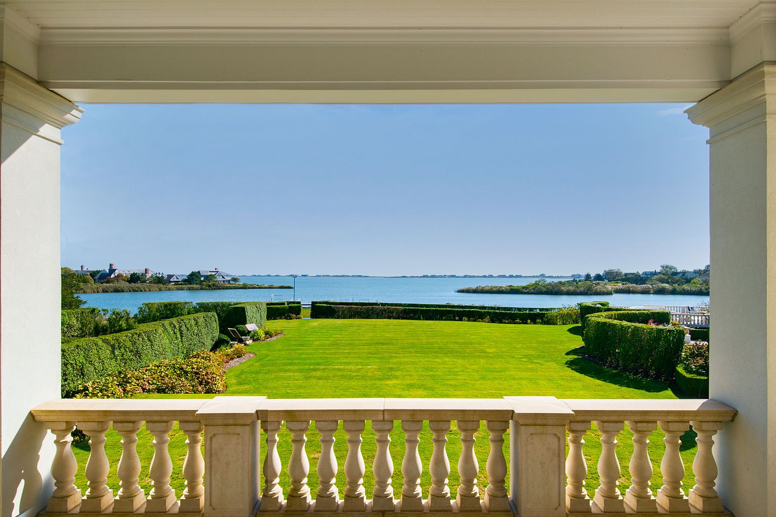 This image showcases a serene view from a balcony overlooking a lush green lawn that extends to the waterfront. The balcony features a classic balustrade, and the scene is framed by architectural columns, creating a sense of elegance and tranquility. The manicured lawn and waterfront view enhance the property's appeal, suggesting a luxurious and peaceful living environment.