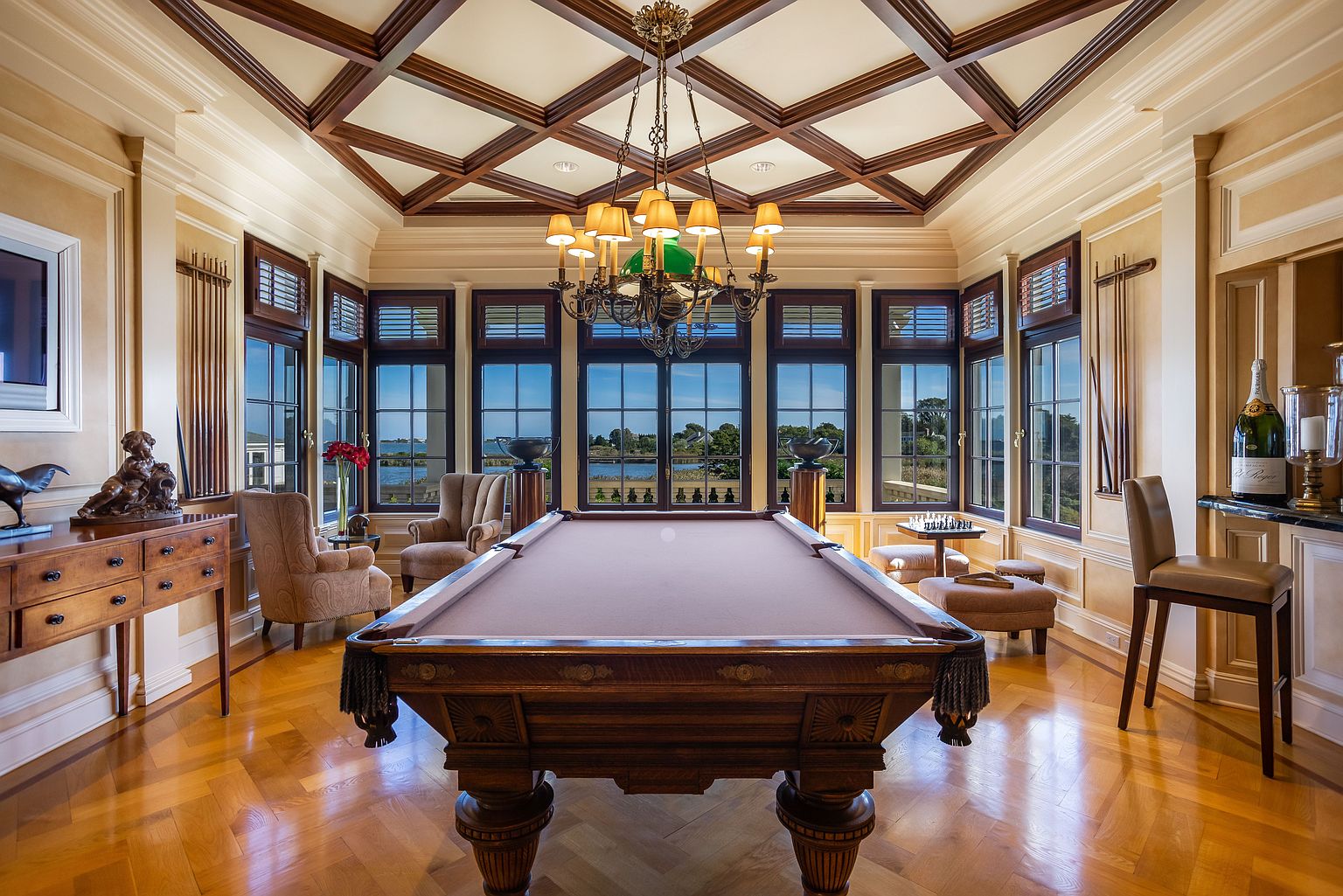 This interior shot showcases a luxurious living room featuring a billiards table as a central element. The room is well-lit with natural light streaming through multiple windows, complemented by an ornate chandelier. The coffered ceiling and hardwood floors add to the room's elegance, creating a sophisticated and inviting space.