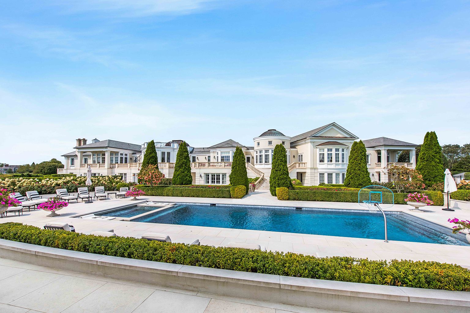 This image showcases a luxurious backyard featuring a large swimming pool with a basketball hoop, surrounded by meticulously manicured hedges and landscaping. The grand mansion serves as a stunning backdrop, emphasizing the property's opulence and providing an idyllic setting for relaxation and entertainment. The perspective is from a slightly elevated position, offering a comprehensive view of the pool area and the impressive residence.