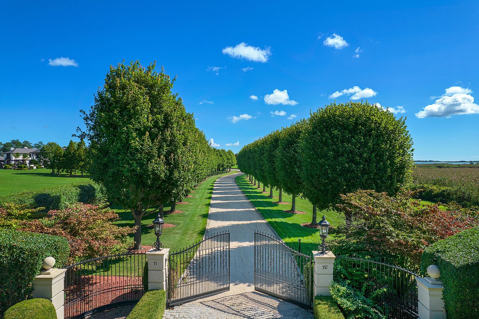 This image showcases a grand entryway to a property, featuring an ornate metal gate flanked by stone pillars with the numbers '71' and '70'. A long, tree-lined driveway stretches into the distance under a bright blue sky with scattered clouds. The landscape is meticulously manicured with lush greenery and flowering bushes, creating an impressive and inviting approach to the estate.
