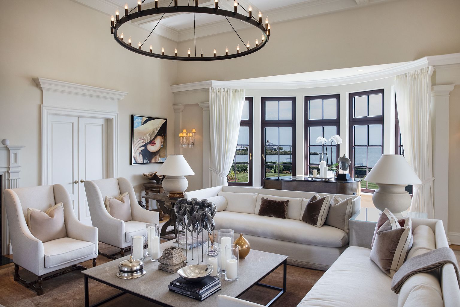 This is a luxurious living room featuring a neutral color palette with white sofas and armchairs, complemented by brown accent pillows. A large, round chandelier hangs from the ceiling, and a bay window offers a view of the water. The room is decorated with various decorative items, including candles, sculptures, and artwork, creating an elegant and inviting atmosphere.