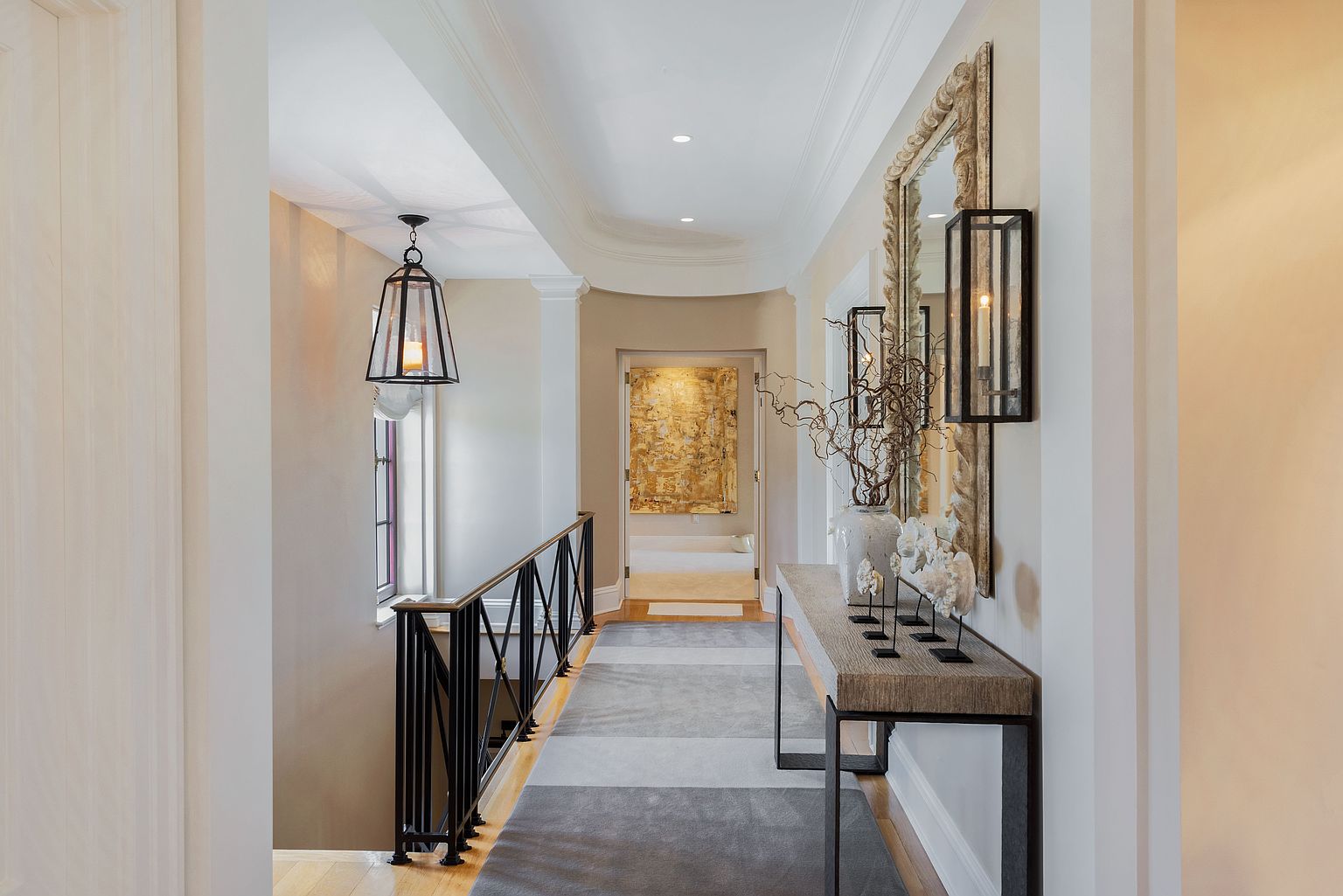 This interior shot showcases a well-lit hallway with a sophisticated design. A black metal railing lines the stairs, while a console table adorned with decorative items and a mirror adds elegance to the space. The neutral color palette and stylish lighting fixtures create a welcoming and upscale atmosphere.