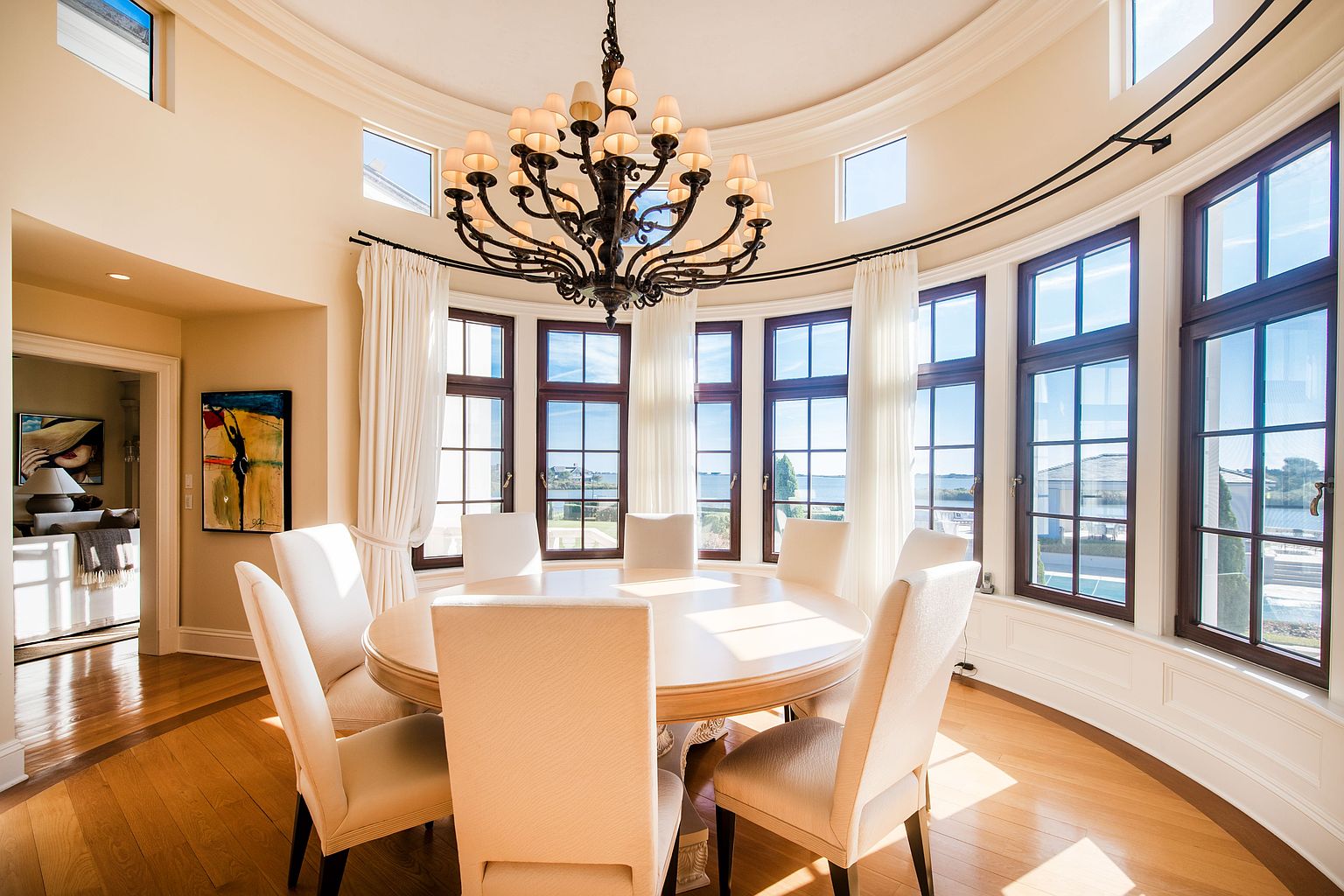 This is an interior shot of a dining room featuring a round table with six chairs, illuminated by a large, ornate chandelier. The room has a curved wall with multiple windows offering a view of the water, and the hardwood floors add warmth to the space. The overall impression is elegant and bright.