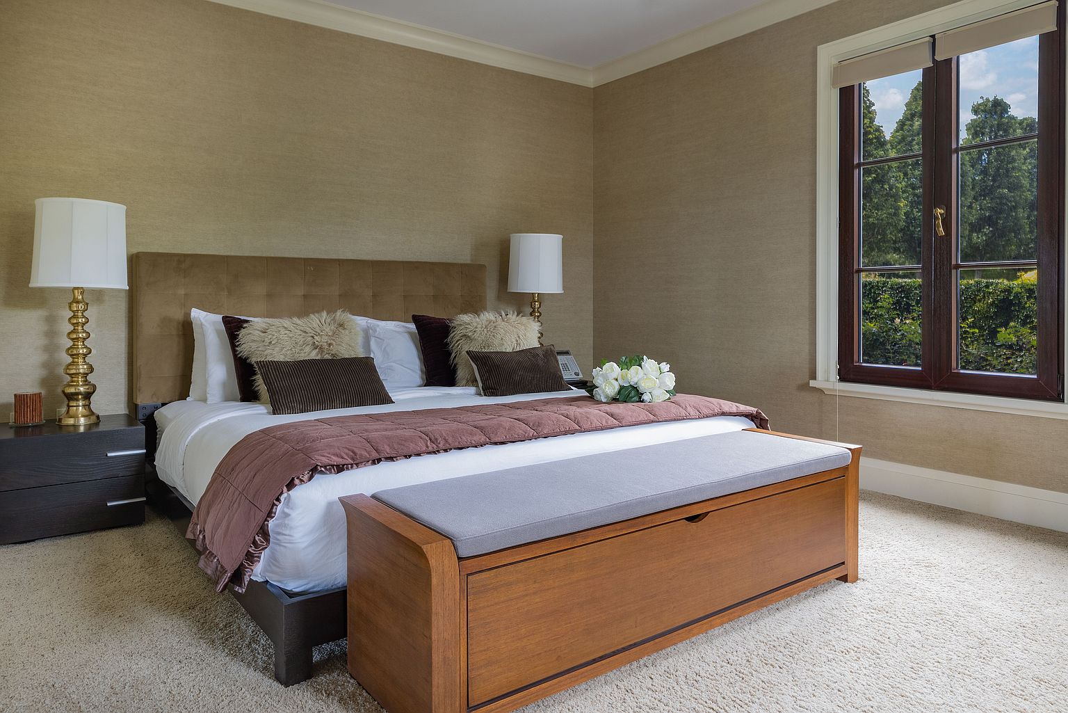 This is a well-appointed primary bedroom featuring a plush bed with a tufted headboard, complemented by decorative pillows and a brown throw. A wooden storage bench sits at the foot of the bed, and a window provides natural light. The room is decorated in neutral tones, creating a warm and inviting atmosphere.
