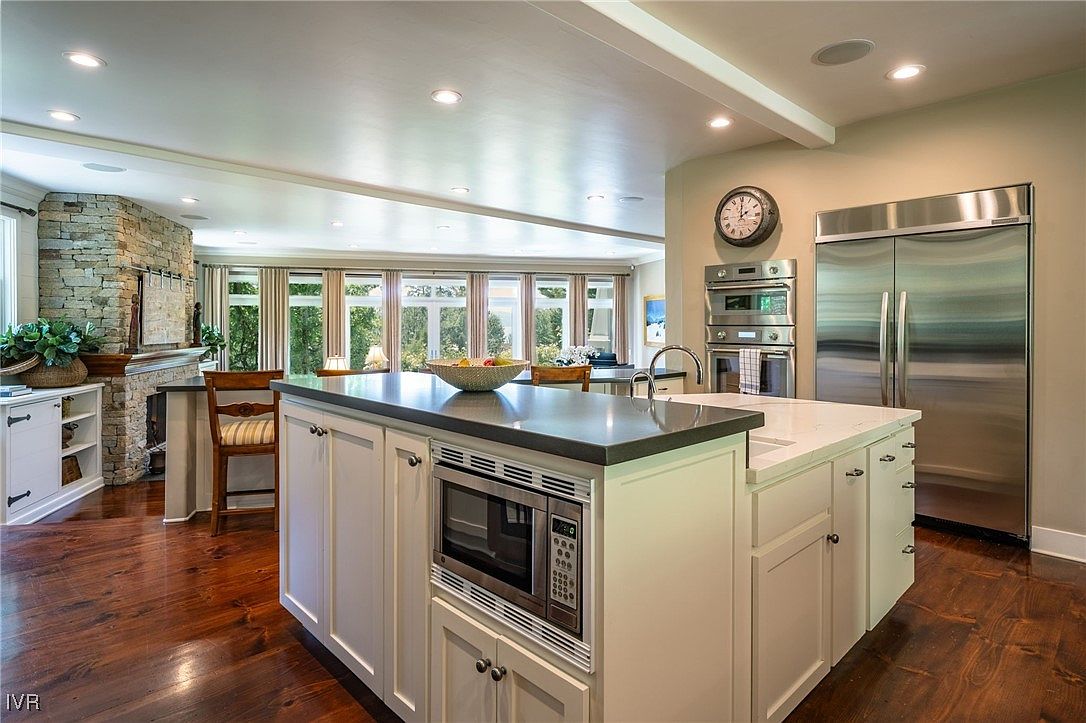 This is a well-lit kitchen featuring a central island with a dark countertop and built-in microwave, complemented by white cabinetry. Stainless steel appliances, including a double-door refrigerator and built-in ovens, add a modern touch. The kitchen opens to a dining area with large windows, providing ample natural light and a view of the outdoors.