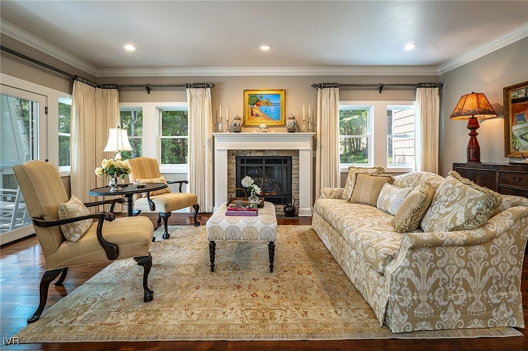 This is a well-decorated living room featuring a fireplace, a patterned sofa, and two armchairs arranged around a central ottoman and rug. The room is illuminated by natural light from the windows and complemented by table lamps, creating a warm and inviting atmosphere. The hardwood floors and neutral wall colors enhance the room's elegance and comfort.