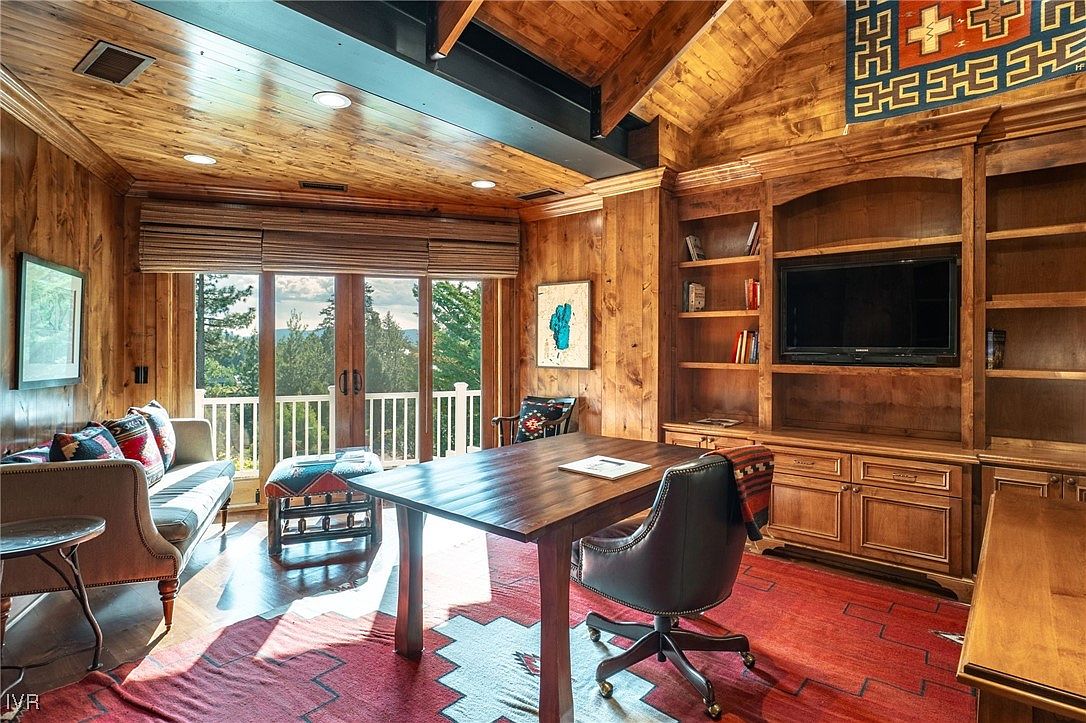 This is a warm and inviting home office featuring wood-paneled walls and ceiling, creating a cozy and sophisticated atmosphere. The room includes a built-in bookcase with a television, a large wooden desk, and comfortable seating, suggesting a functional and stylish workspace. Natural light streams in through the windows, enhancing the room's appeal.
