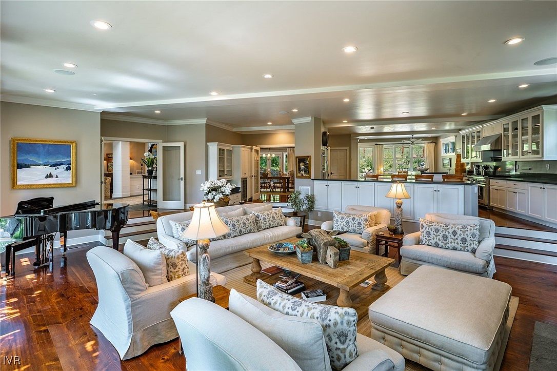 This is an interior shot of a luxurious living room that seamlessly transitions into a kitchen area. The living room features comfortable, neutral-toned sofas and armchairs arranged around a rustic wooden coffee table, with a grand piano visible in the background. The kitchen boasts white cabinetry and modern appliances, creating an open and inviting space perfect for entertaining.