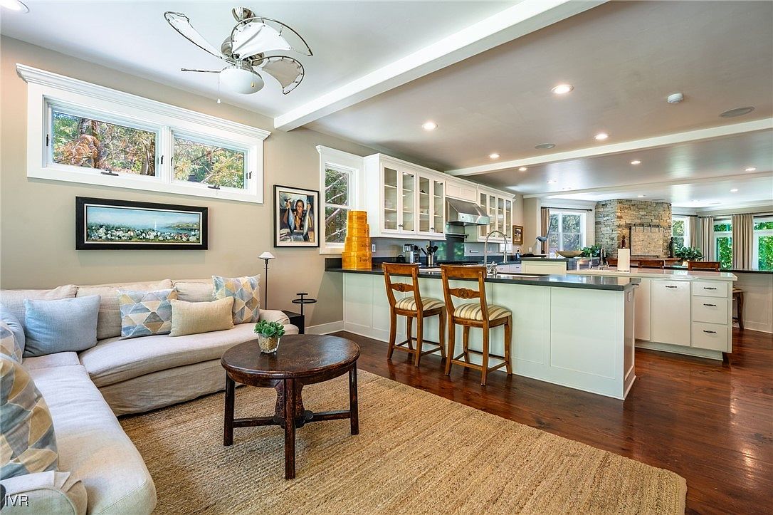 This interior shot showcases a combined living room and kitchen area. The kitchen features white cabinetry, a dark countertop island with bar seating, and stainless steel appliances. The living room area includes a comfortable sectional sofa, a round wooden coffee table, and a large area rug, creating a cozy and inviting atmosphere.