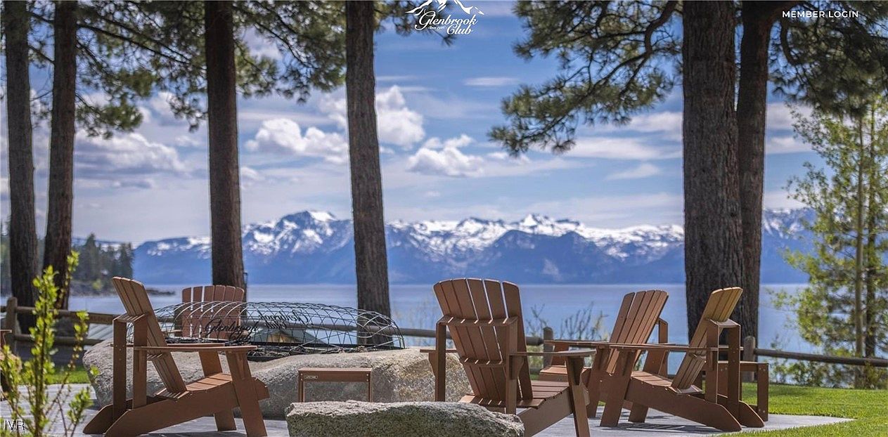 This image showcases an inviting outdoor patio area with Adirondack chairs arranged around a fire pit, set against a stunning backdrop of a lake and snow-capped mountains. The scene is framed by mature trees, creating a sense of privacy and tranquility. The overall impression is one of luxury and relaxation, perfect for enjoying the natural beauty of the surroundings.