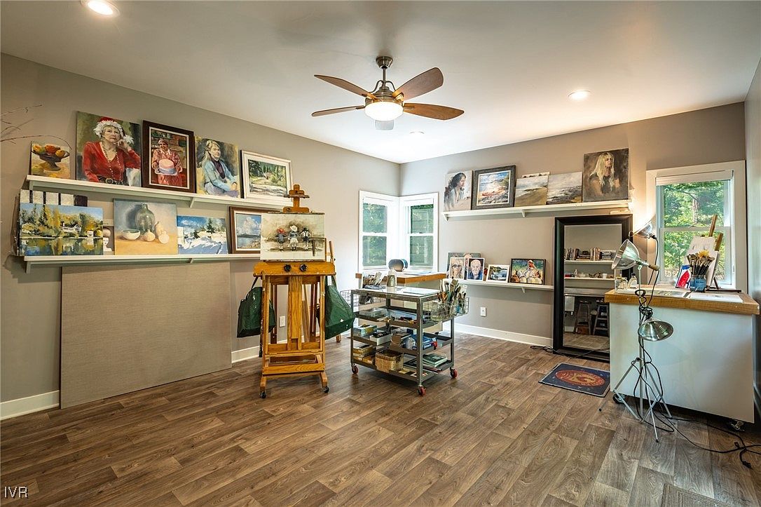 This is an interior shot of an artist's studio or office space. The room features hardwood floors, neutral-colored walls, and a variety of artwork displayed on shelves. An easel stands prominently in the center, and a rolling cart holds art supplies, suggesting a creative and functional workspace.