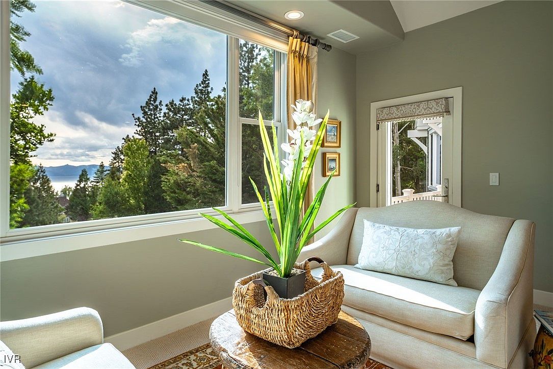 This interior shot showcases a cozy living room area with a focus on natural light and serene views. A comfortable beige sofa with a decorative pillow sits near a large window overlooking a lush landscape with trees and a glimpse of a lake. A decorative plant in a woven basket sits on a wooden table, adding a touch of nature to the indoor space.