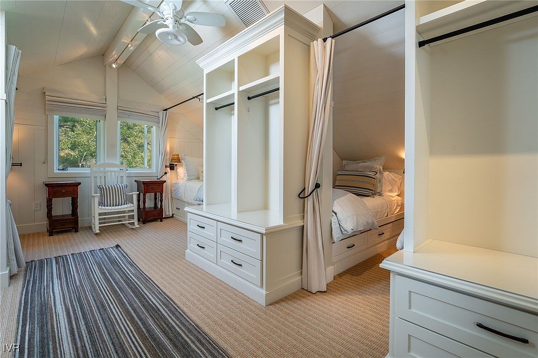 This is a charming guest bedroom featuring two built-in twin beds separated by a central wardrobe unit with drawers. The room has a cozy, attic-like feel with sloped ceilings and white wood paneling. Natural light streams in through the windows, complemented by soft bedside lighting, creating a serene and inviting space.