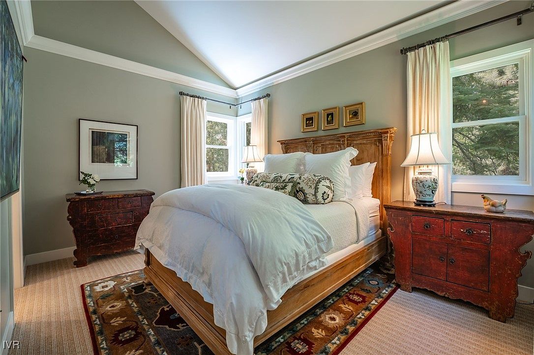 This is a well-lit primary bedroom featuring a wooden bed frame with white bedding and decorative pillows. The room includes two wooden side tables, each with a lamp, and framed artwork on the walls. A patterned rug adds warmth to the carpeted floor, and natural light streams in through the windows, creating a cozy and inviting atmosphere.