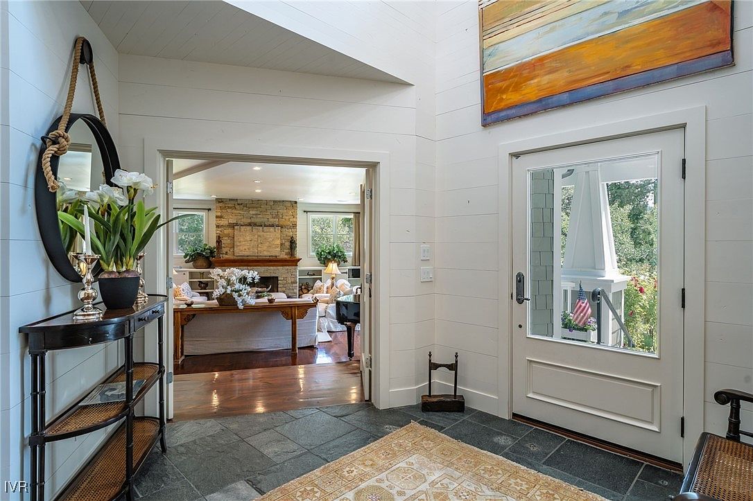This interior shot showcases a well-lit entryway with white shiplap walls and a glimpse into a cozy living room. A dark wood console table with a round mirror and floral arrangement adds a touch of elegance, while a patterned rug on the slate tile floor provides visual interest. The open doorway invites viewers into the adjacent living space, creating a sense of flow and connection.