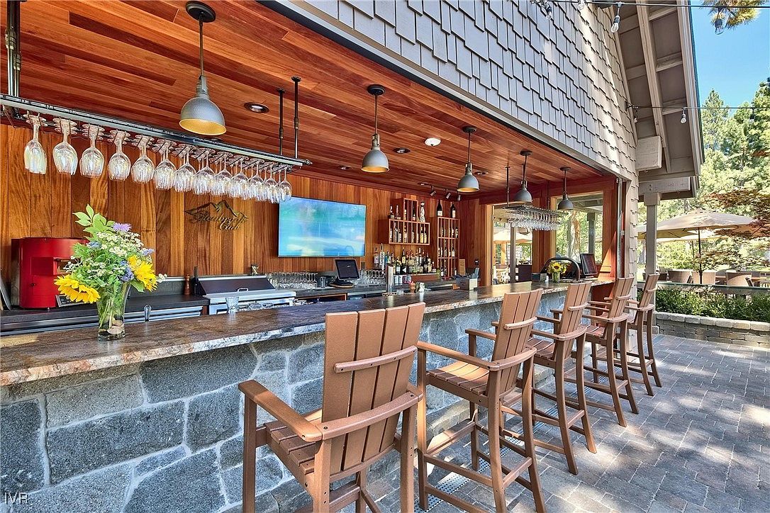 This image showcases an outdoor bar area, featuring a stone-clad bar counter with wooden bar stools. Overhead, pendant lights illuminate the space, and a wine glass rack is visible. The patio is paved with stone tiles, and greenery surrounds the area, suggesting a relaxed and upscale outdoor entertainment space.