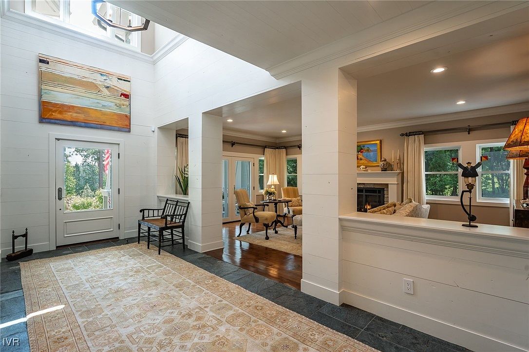 This interior shot showcases a bright and airy hallway leading into a living space. The hallway features a neutral-toned rug, white shiplap walls, and a high ceiling with a skylight, creating an open and inviting atmosphere. The transition into the living room reveals hardwood floors and comfortable seating, suggesting a seamless flow between spaces.