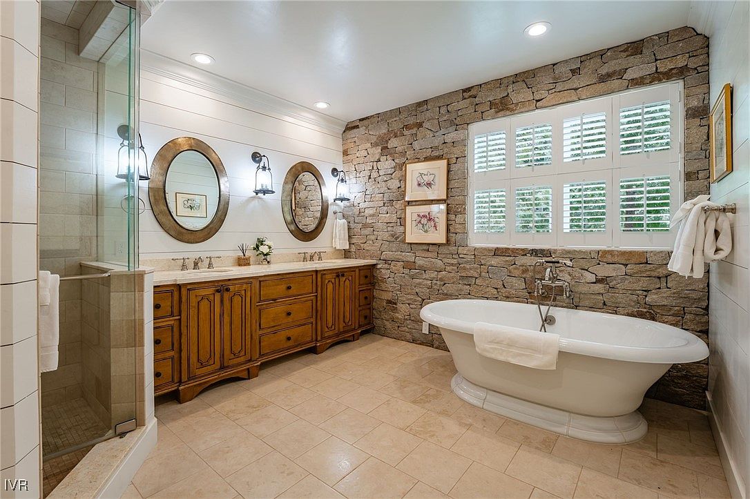 This is a luxurious primary bathroom featuring a freestanding bathtub set against a stone accent wall with a window. The room includes a double vanity with wooden cabinetry, oval mirrors, and sconce lighting. A glass-enclosed shower is visible to the left, and the overall design exudes a spa-like ambiance with a blend of rustic and elegant elements.